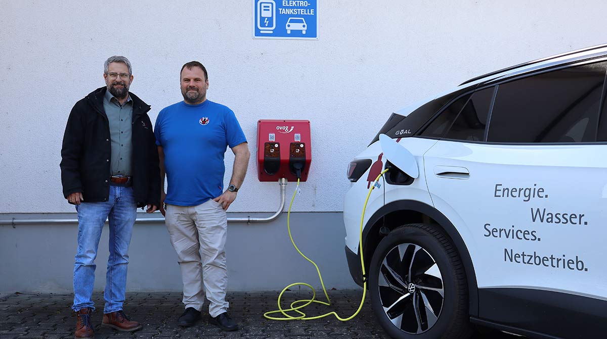 Dirk Schneider und Christian Weber stehen an der vereinseigenen Sporthalle neben der neuen Wallbox.