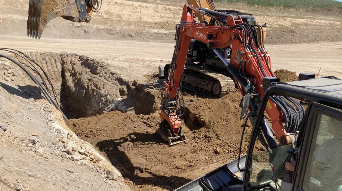 Zwei Bagger heben eine große Baugrube aus, während freigelegte Kabel sichtbar sind. Ein Arbeiter steuert die Maschine.