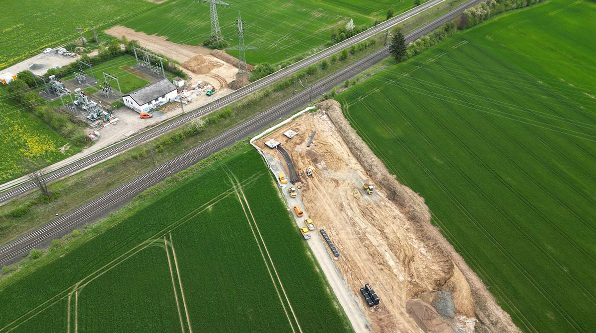 Eine Luftaufnahme zeigt die Baustelle neben den Bahngleisen und dem Umspannwerk, umgeben von grünen Feldern.